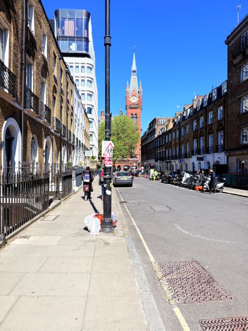 A street scene in Brixton showing terraced residential buildings with brick facades and white window frames on both sides of the road, under a clear blue sky. The sidewalk features a black metal fence in front of each property, with a few pedestrians walking and carrying bags. Parked motorcycles and small cars are aligned along the curb, with some vehicles further down the street. In the background, a prominent red brick clock tower with a pointed spire rises above the other buildings, while a modern curved glass office structure is visible on the left. A black street lamp is positioned in the foreground, partially obscuring the view of the loading and unloading activities associated with house removals and furniture transport services. The scene captures a typical urban environment suitable for home relocation and moving logistics, with Brixton Removals potentially operating in this area.