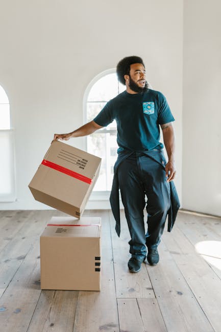 A professional mover from Brixton Removals is inside a bright, empty room with white walls and light wooden flooring, preparing for a home relocation. He is wearing a dark blue uniform with a company logo on the chest and is holding a medium-sized cardboard box wrapped with red tape in his right hand. Two additional cardboard boxes, one with black tape on the side, are placed on the floor nearby, indicating packing and moving preparations. The room features a large arched window with natural light streaming in, illuminating the scene. The mover's posture suggests he is about to load the box onto a trolley or transport it through the doorway for furniture transport or loading into a removal van. The setting captures the early stages of packing and furniture transport within a residential environment, aligned with [PAGE_TITLE] and the services offered by [COMPANY_NAME] for efficient house removals in Brixton.